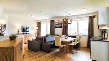 A modern hotel room with a wooden floor, a dining area with a table and chairs, a sofa, a TV, and a kitchen area with a refrigerator and a coffee machine.