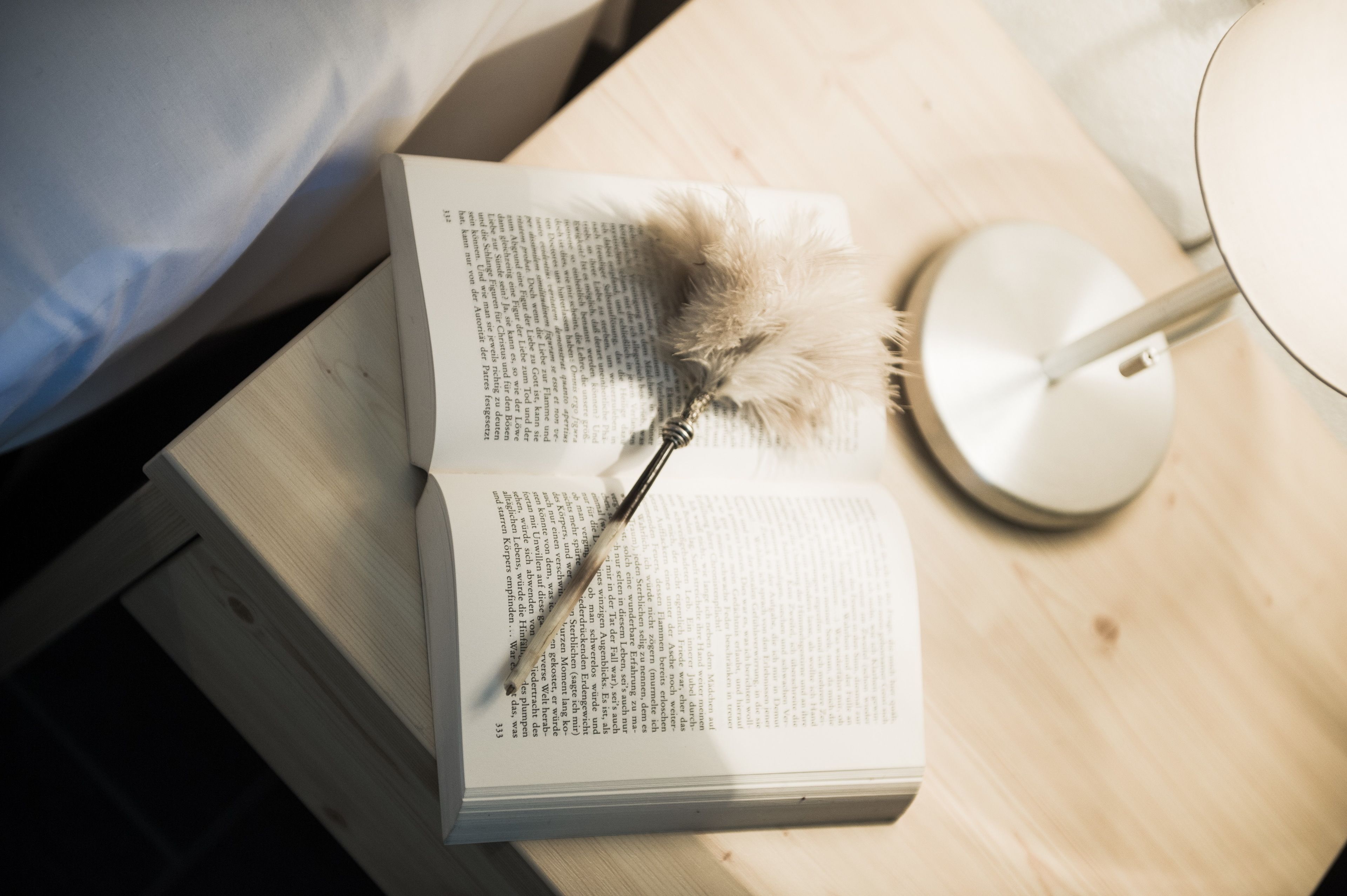 Ein offenes Buch auf einer Holztischplatte, dazu eine Federkiele und eine Lampe in der Nähe.