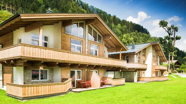 Een houten huis met twee verdiepingen, een balkon, stenen decoraties en uitzicht op een beboste helling.