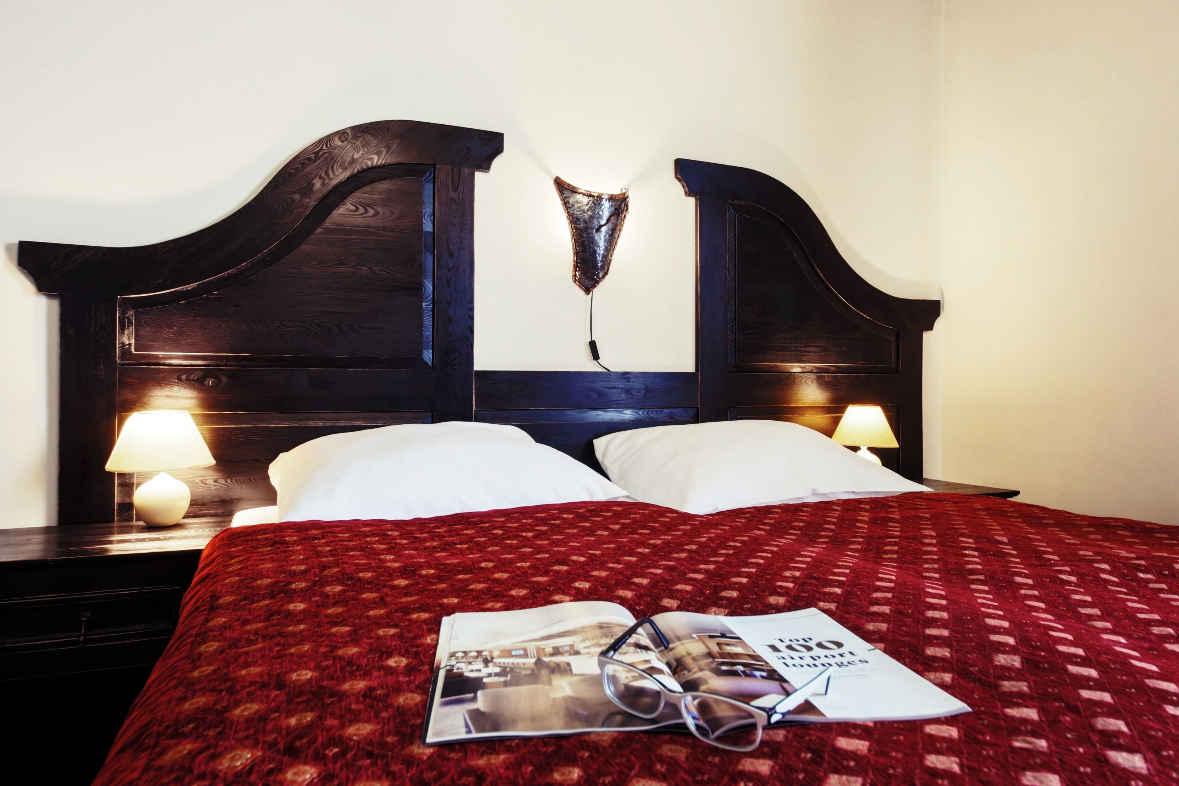 Una cama cuidadosamente tendida con una colcha roja estampada, dos almohadas blancas y una revista con gafas encima.
