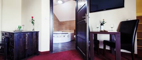 Habitación de hotel con alfombra roja, un armario de madera, un televisor, una mesa con copas de vino y una bañera.