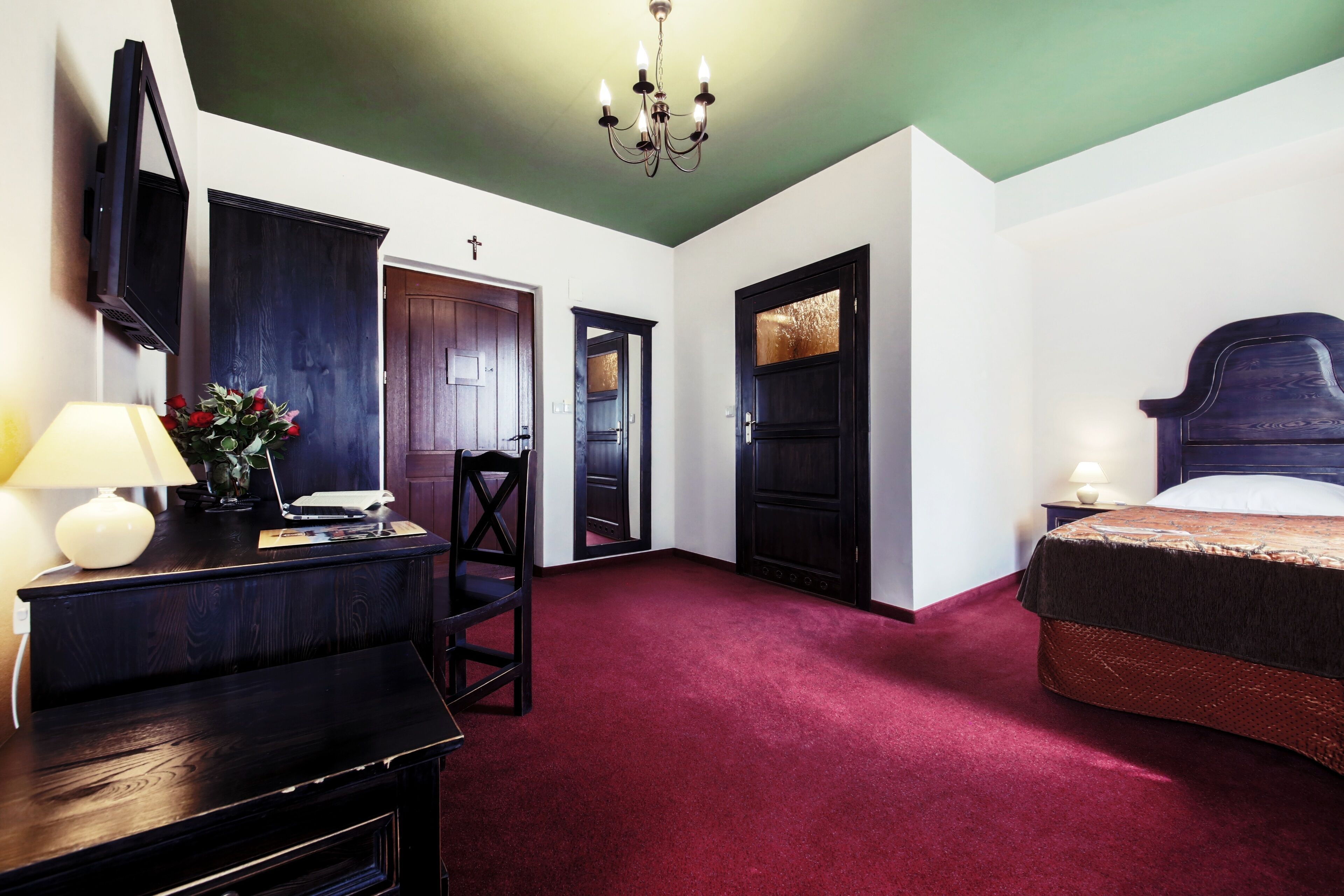 A room with a dark wooden desk, a chair, a bed with a headboard, a nightstand with a lamp, and a wall-mounted TV.