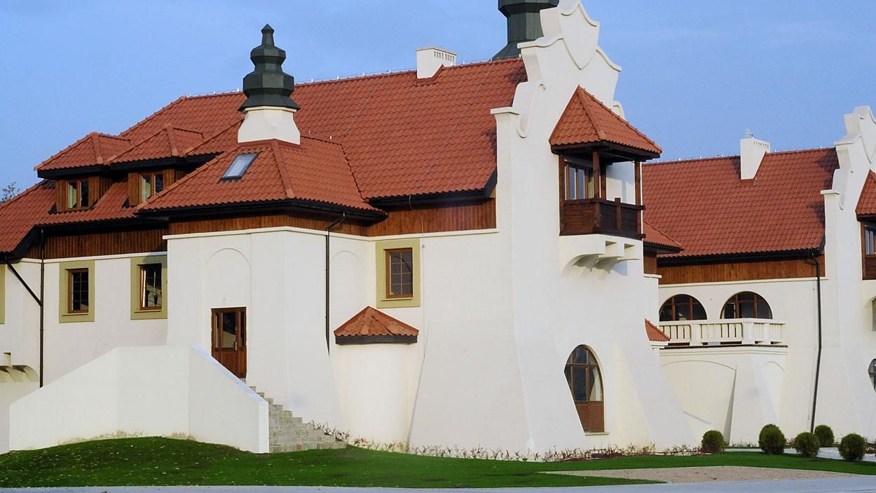 A white building with red-tiled roofs and multiple chimneys. It has a staircase leading to a balcony and several windows.