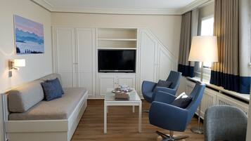 A modern hotel room with a sofa, two armchairs, a coffee table, a flat-screen TV, and a lamp.