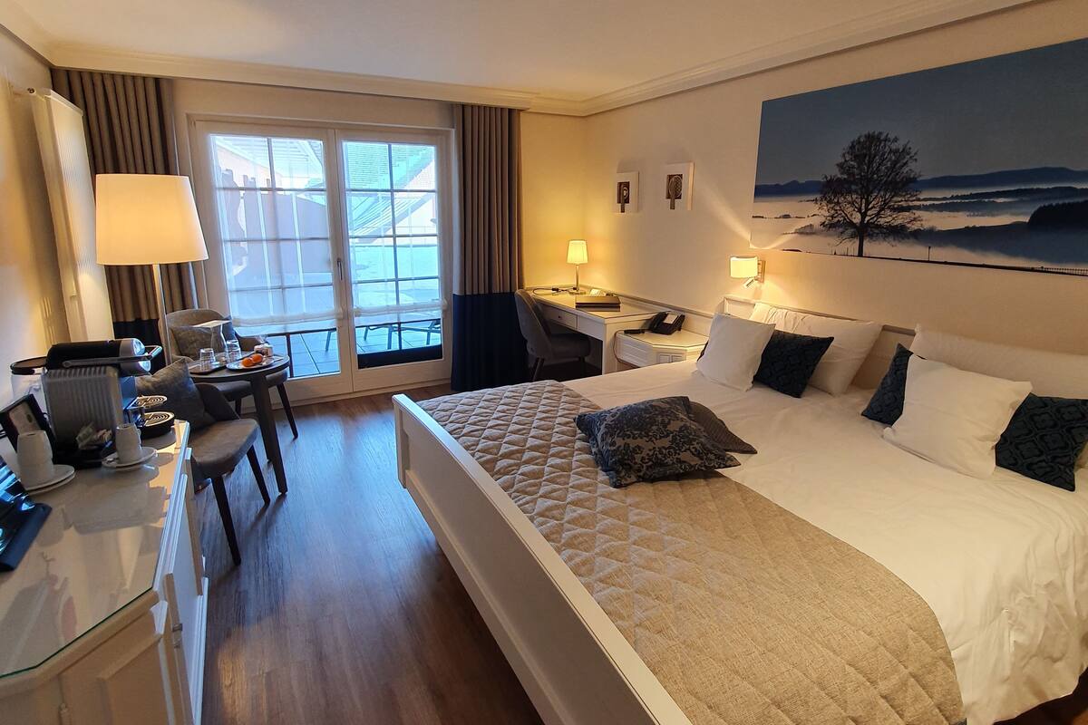 A hotel room with a large bed, a desk with a lamp, a chair, a dining table with breakfast items, and a painting on the wall.