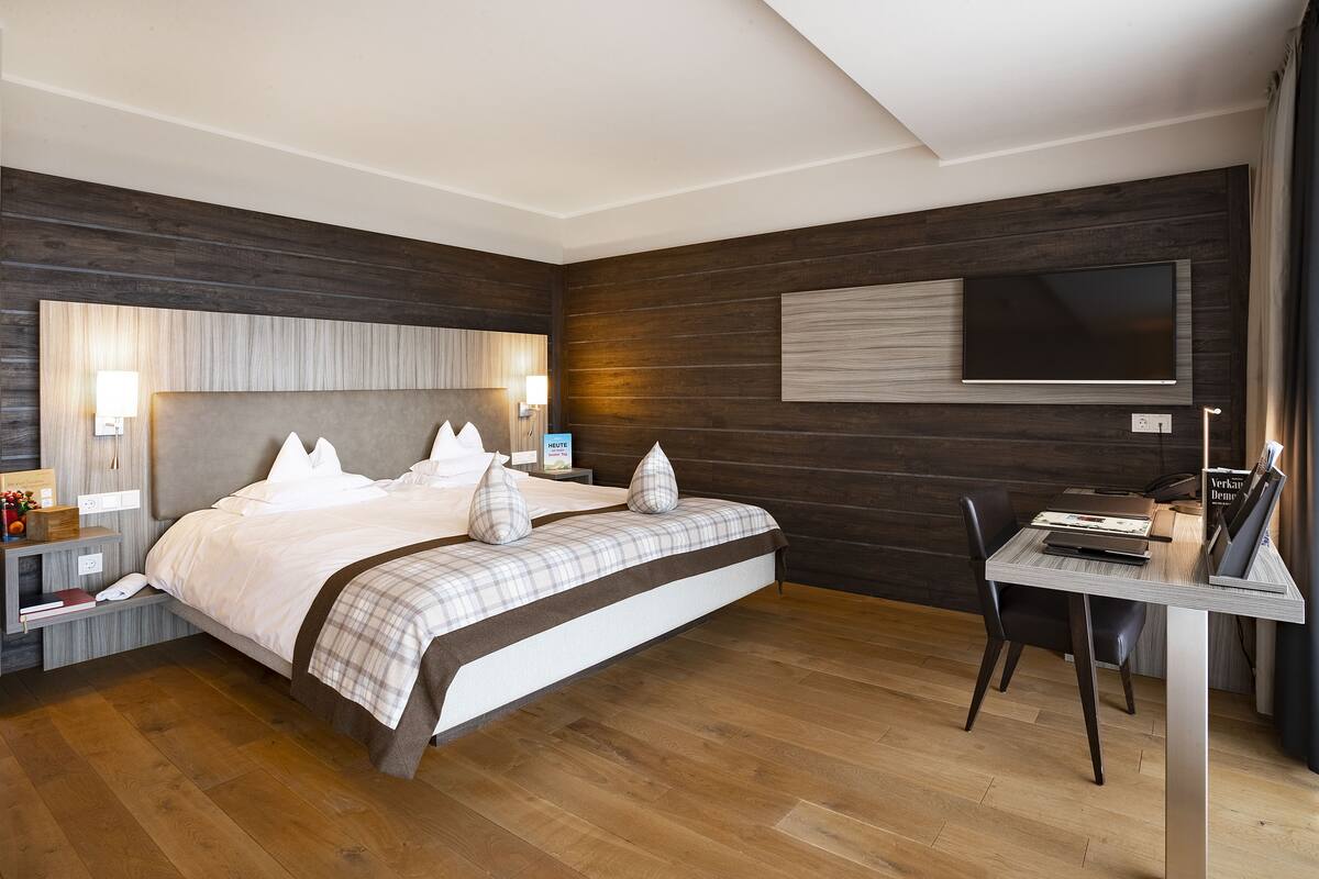 A modern hotel room with a large bed, bedside tables, a desk with a chair, a flat-screen TV, and wooden paneling on the walls.