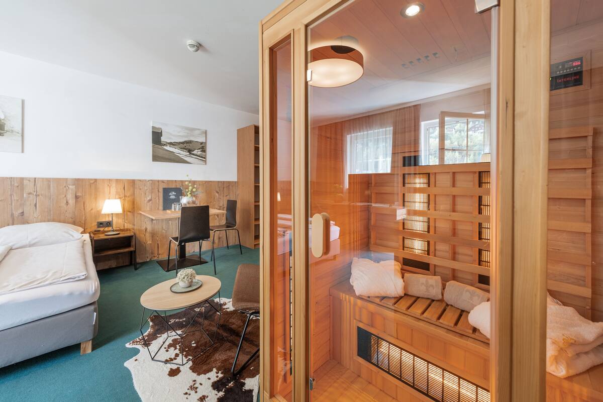 A modern hotel room with a wooden interior, a cozy seating area with a sofa and a small table, a wooden sauna area with towels and benches, and a large window providing natural light.