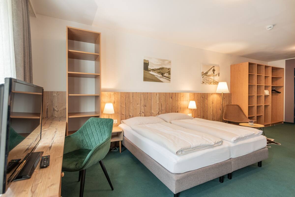 A hotel room with a bed, a desk with a chair, a TV, a wardrobe, and a lamp.