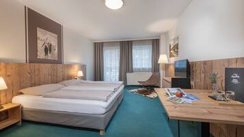 Ein Hotelzimmer mit einem großen Bett, einem Schreibtisch mit Lampe, einem Sessel, einem Fernseher und einem Fenster mit Vorhängen.