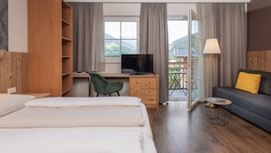 A hotel room with a bed, a desk with a computer, a TV, a sofa with a yellow pillow, and a balcony with a view of the mountains. - AlpenParks Hotel Montana Matrei (Matrei in Osttirol)