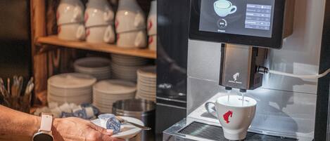 Una cafetera con pantalla digital, una taza y una persona sosteniendo un plato con una cuchara.