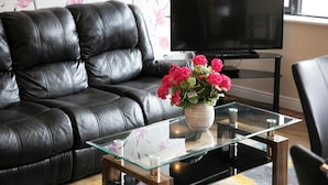A living room with a black leather sofa, a glass coffee table with a vase of flowers, a flat-screen TV on a stand, and a window with a floral wallpaper. - Arch House Apartments (Athlone)