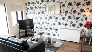 A modern living room with a black leather sofa, a glass coffee table, a flat-screen TV, and a floral-patterned wall. - Arch House Apartments (Athlone)