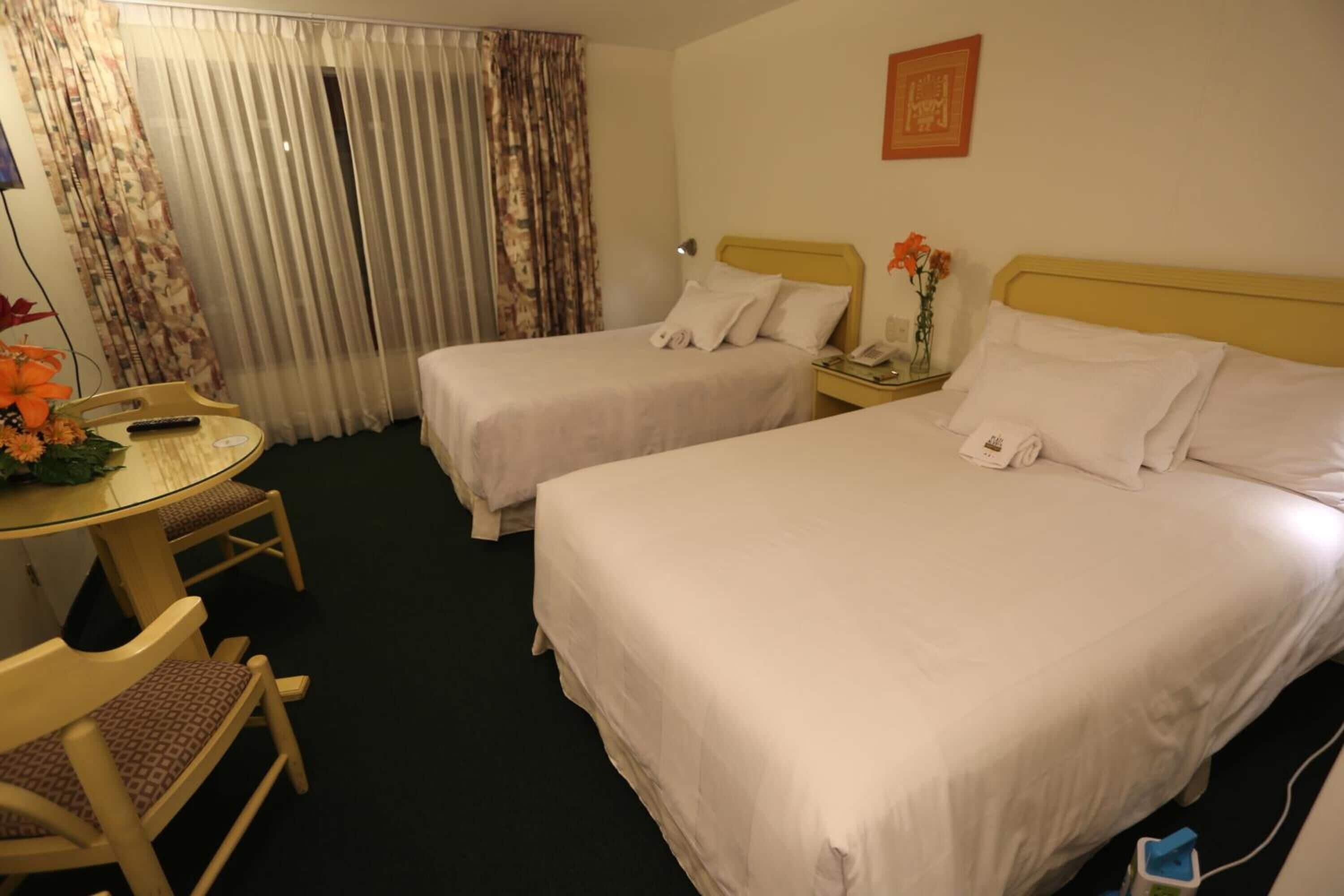 Une chambre d’hôtel avec deux lits, une petite table avec des chaises, un téléphone et un vase avec des fleurs.