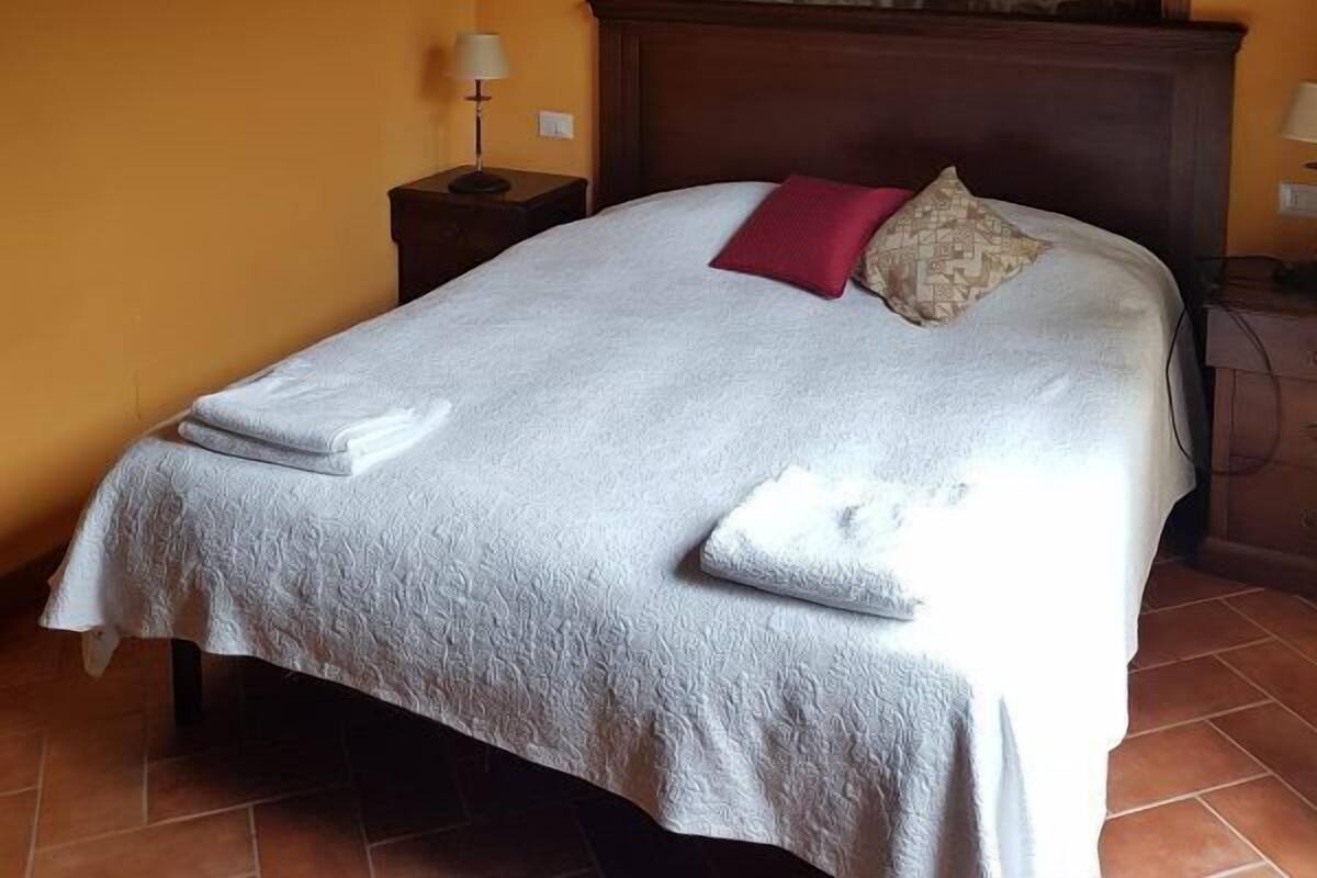 Une chambre avec un mur en pierre, une tête de lit en bois, un lit recouvert d’une courtepointe blanche et agrémenté de deux oreillers, ainsi que deux tables de chevet avec des lampes.