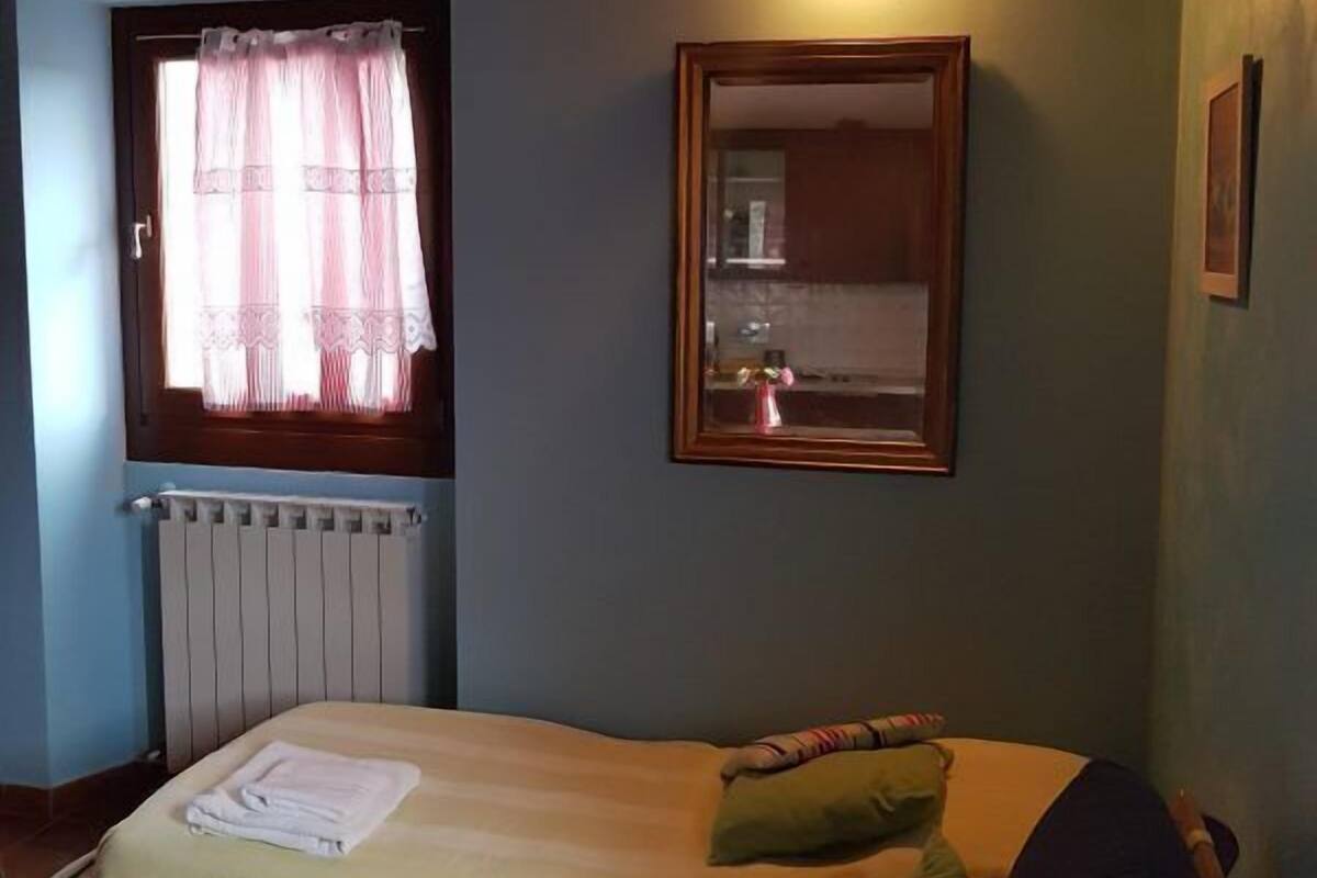Une pièce avec un lit, une fenêtre avec des rideaux, un radiateur, un miroir et un plafond en bois.
