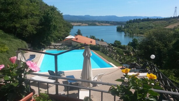 Une piscine avec vue sur un lac et des montagnes, entourée de verdure et dotée d’une terrasse en bois avec des chaises longues.