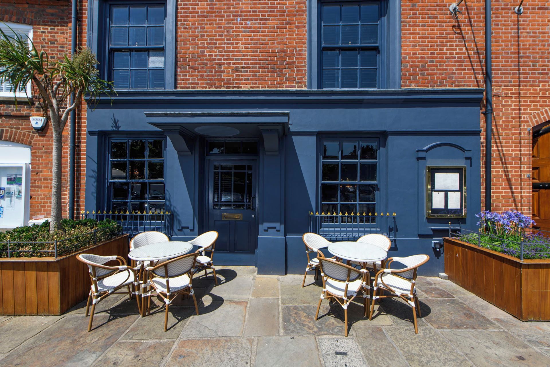 A building with a dark blue facade, multiple windows, and a door. There are outdoor wicker chairs and a table in front.