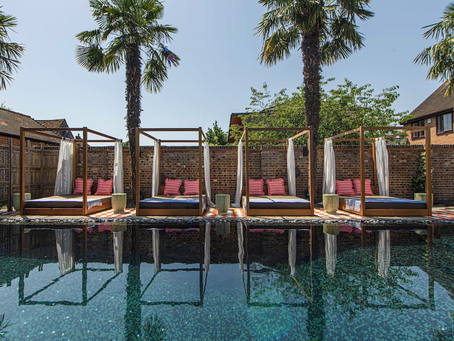 A poolside area with three wooden beds, each with a canopy and pillows, set against a brick wall with palm trees.