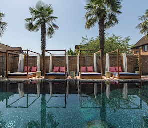 Un área junto a la piscina con tres camas de madera, cada una con dosel y almohadones, frente a una pared de ladrillo con palmeras.