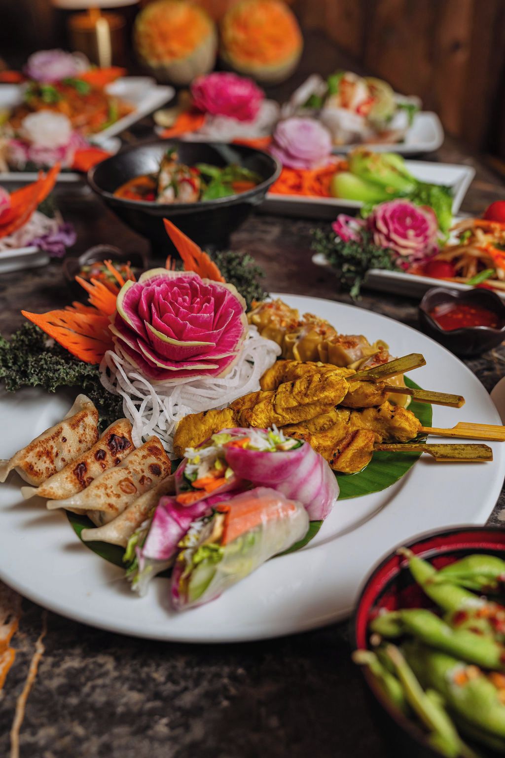 Verschiedene Gerichte, darunter Frühlingsrollen, gegrilltes Fleisch und Spieße, auf einem weißen Teller mit einer Zierblume angerichtet.