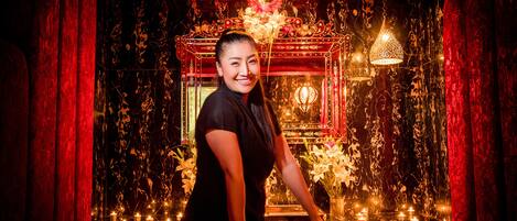 A woman sitting on a bed in a dimly lit room with red curtains and a decorative mirror.