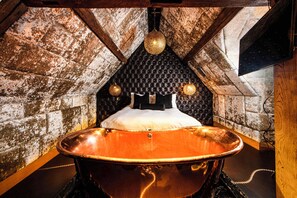 A room with a large copper bathtub, a black tufted headboard, and a stone wall. - Crazy Bear Hotel-Beaconsfield (Beaconsfield)