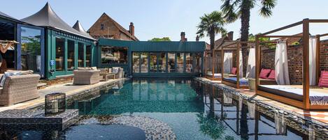 A swimming pool with lounge areas, a covered seating area, and a brick building in the background.