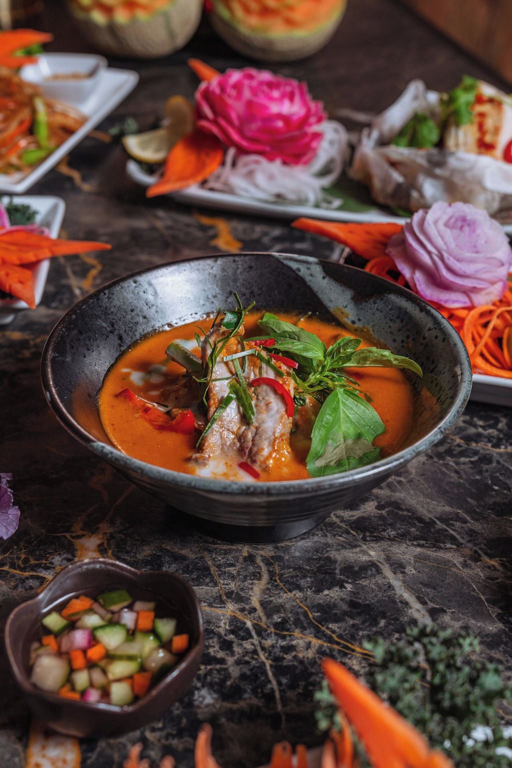 Un tazón de sopa con verduras y hierbas, rodeado de distintos platos y guarniciones sobre una mesa de mármol.