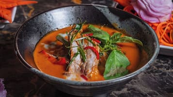 Un tazón de sopa con verduras y hierbas, rodeado de distintos platos y guarniciones sobre una mesa de mármol.
