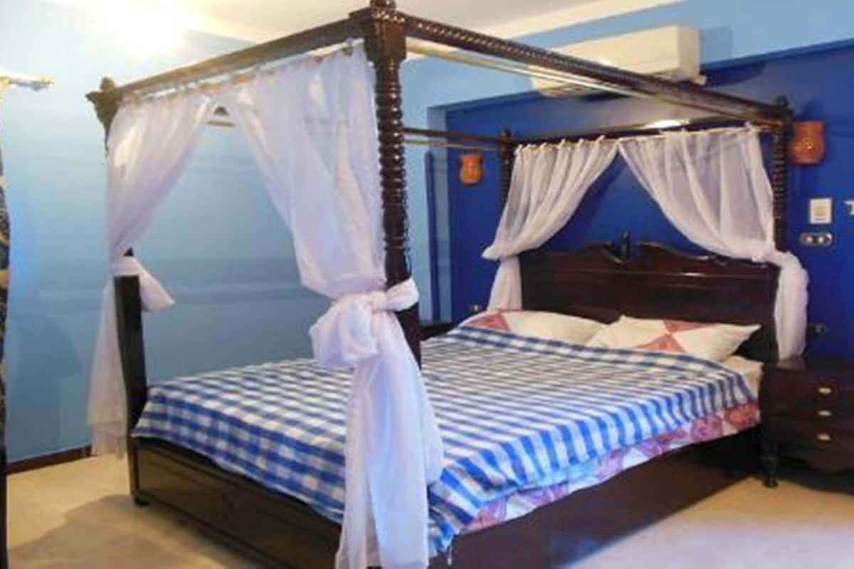 Una cama con dosel y cuatro columnas, con edredón a cuadros, cabecera de madera y una mesita de noche con lámpara.