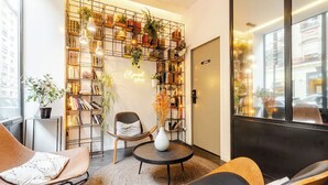 A cozy reading nook with a bookshelf, a comfortable chair, a small table, and a potted plant. - Atypik Hôtel - Urban Style (Clichy)