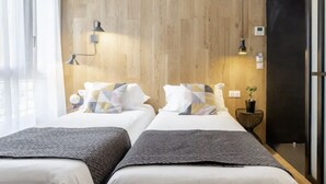 A modern hotel room with two beds, wooden paneling, a small side table with a plant, and a window with sheer curtains. - Atypik Hôtel - Urban Style (Clichy)