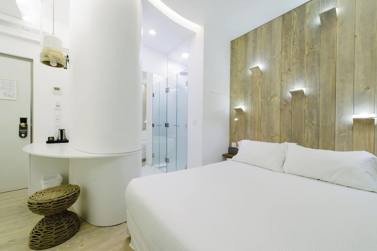Una habitación de hotel moderna con una cama grande, un muro decorativo de madera, una mesa redonda blanca y un box de ducha de vidrio.