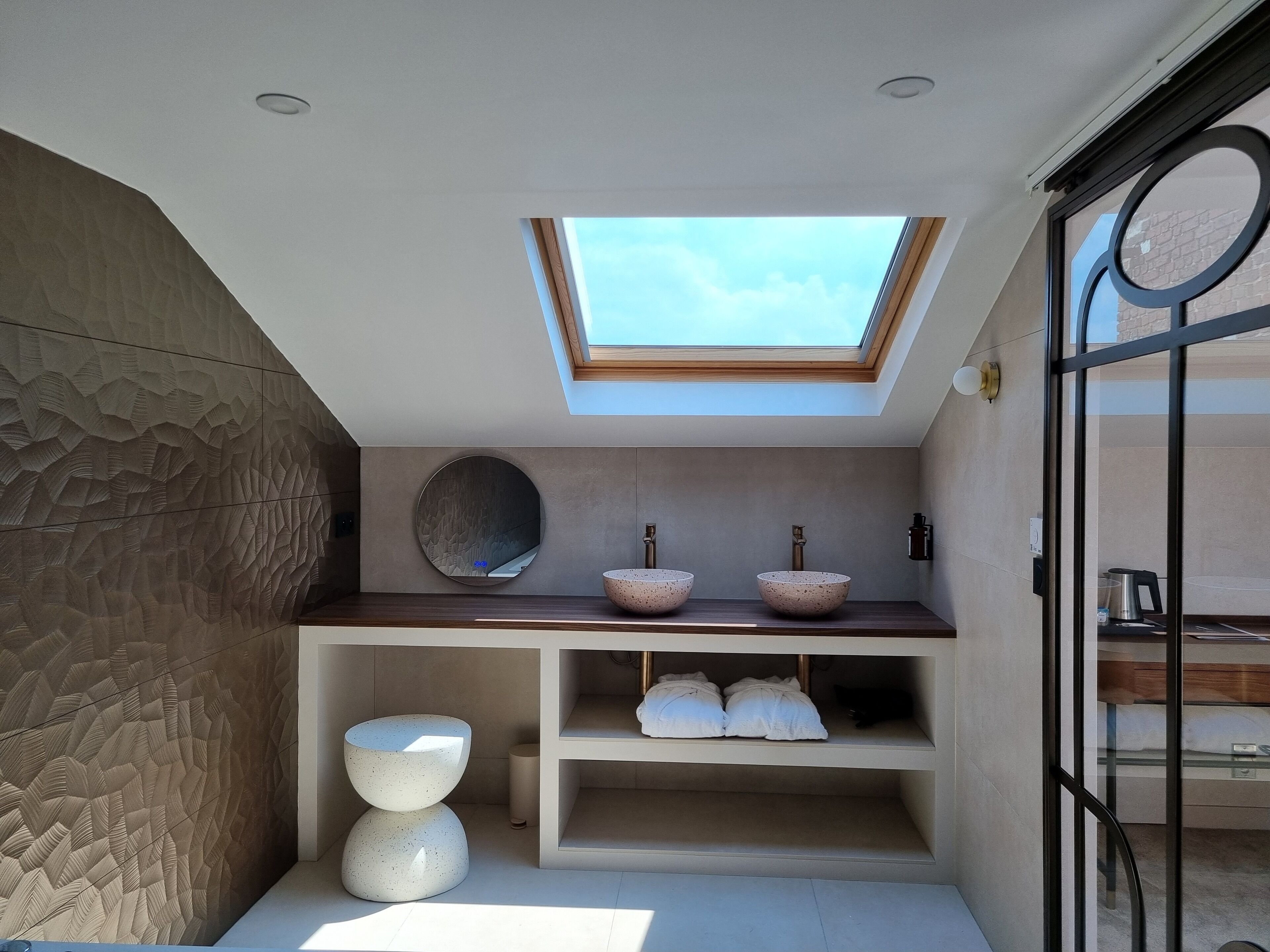 A modern bathroom with a skylight, two vessel sinks, a round mirror, and storage shelves.