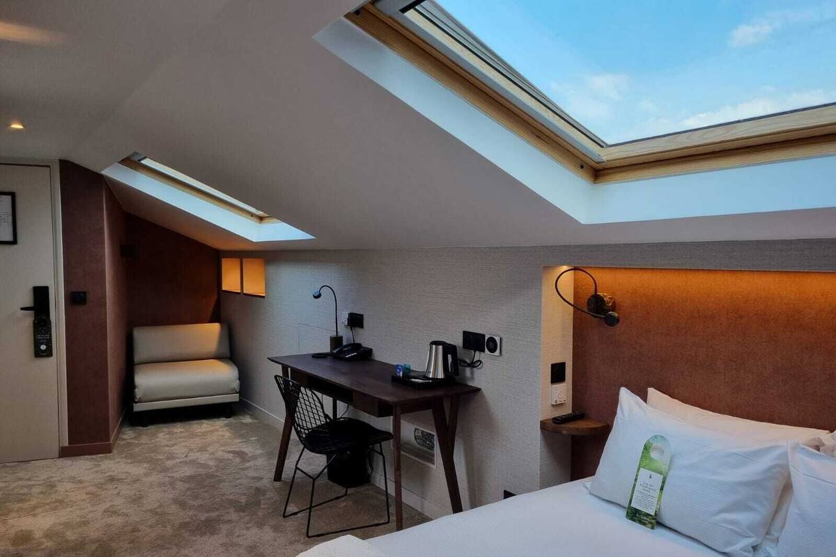 Habitación de hotel con tragaluz, escritorio, silla, cama y mesita de noche con lámpara.