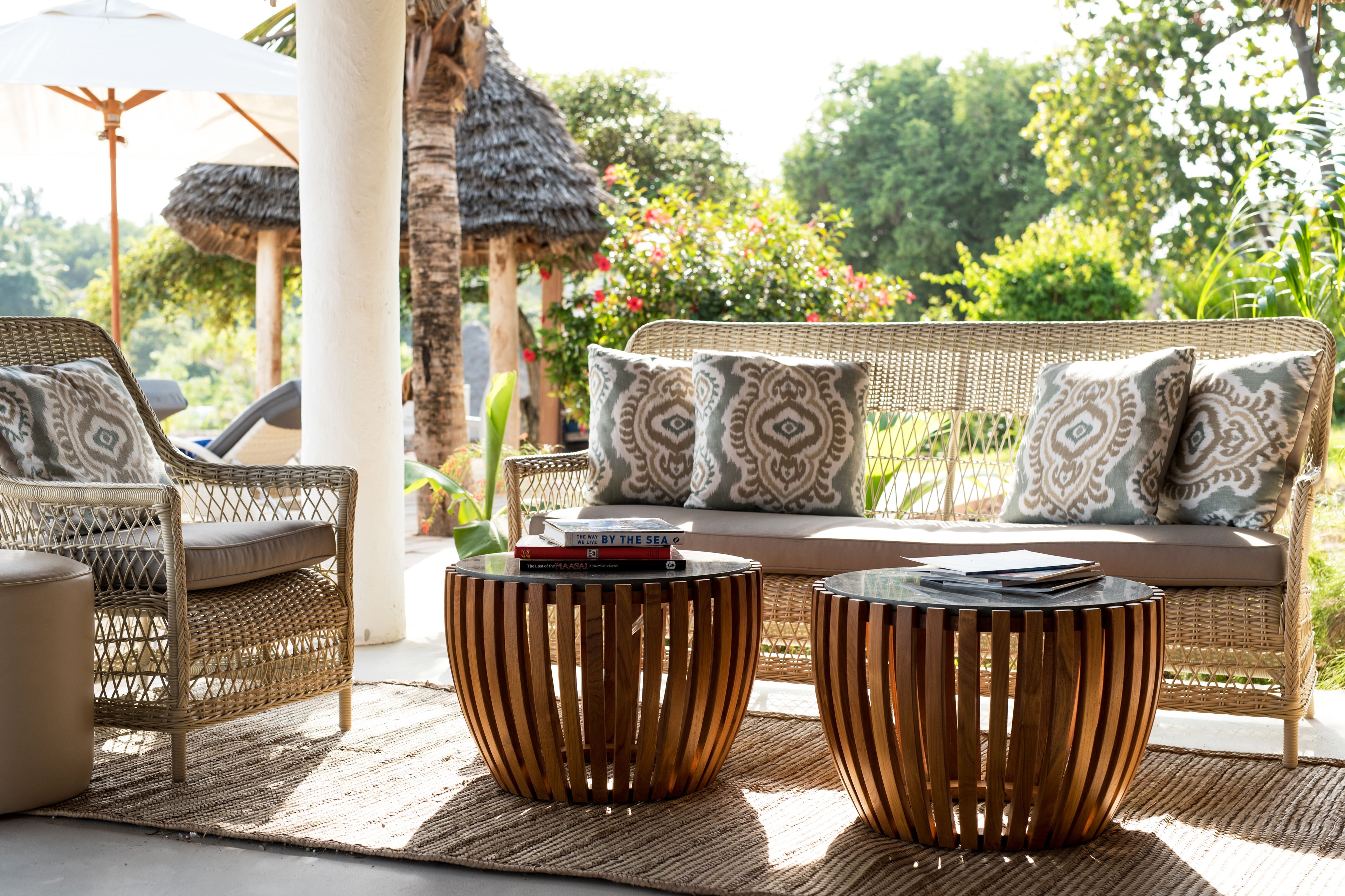 A wicker sofa with patterned cushions, two wooden side tables, and a woven mat on a patio with a thatched roof and lush greenery in the background.