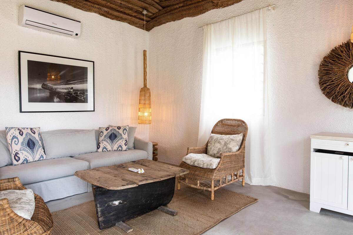 Ein Wohnzimmer mit einer Couch, einem Holztisch, einem Korbstuhl, einem Korb, einer Wandklimaanlage, einem gerahmten Bild, einer Pendelleuchte und einem weißen Schrank.
