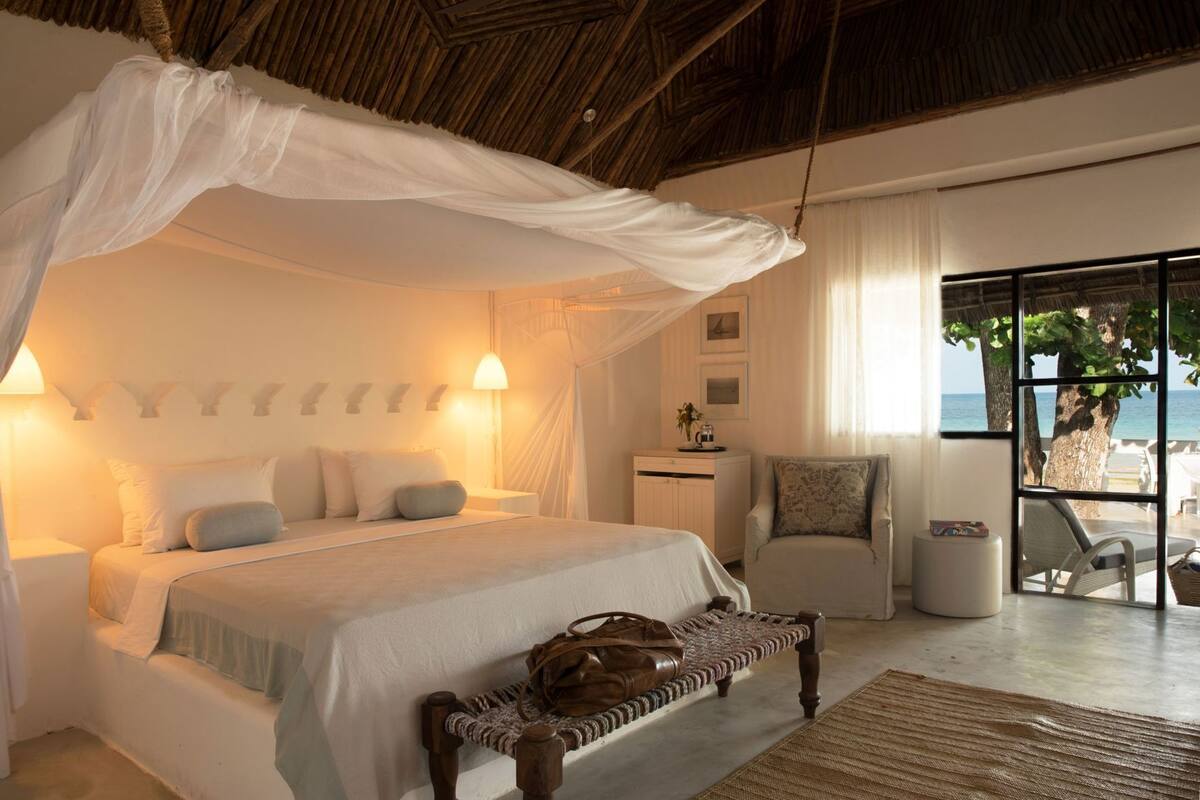 Ein Schlafzimmer mit Baldachinbett, Nachttischlampen, einer Bank mit einer Tasche, einem Nachtkästchen und Blick auf das Meer durch eine Glastür.