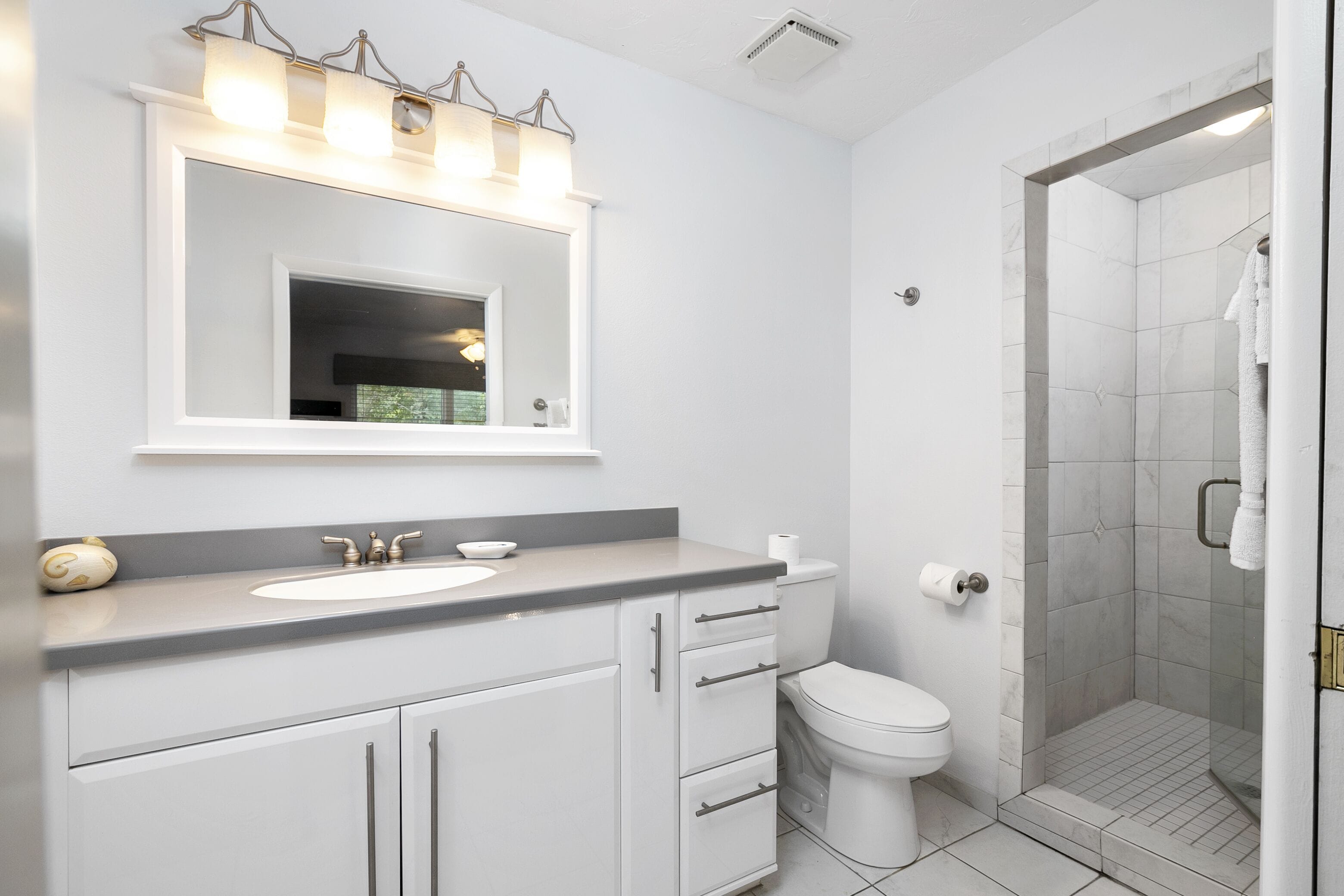 A modern bathroom with a white sink, vanity, and toilet. It includes a shower area with a glass door and a mirror above the sink.