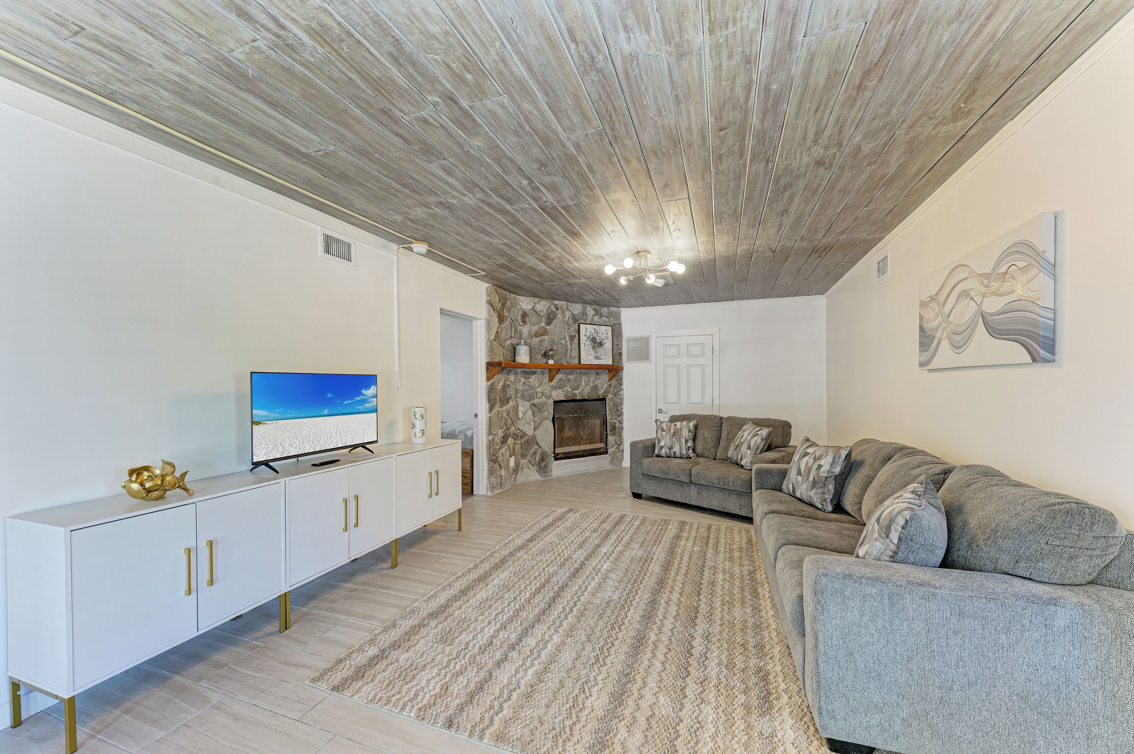 A modern living room with a stone fireplace, a grey sofa, and a flat-screen TV on a white cabinet.