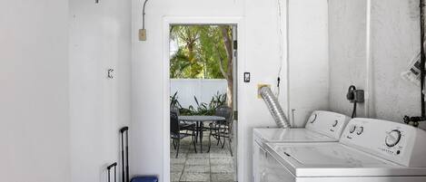 A laundry room with a washing machine and dryer, a small table with chairs, and a door leading to an outdoor area with plants.
