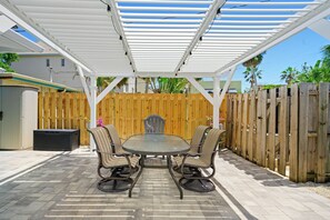 An outdoor patio with a covered table and chairs, a wooden fence, and a house in the background.