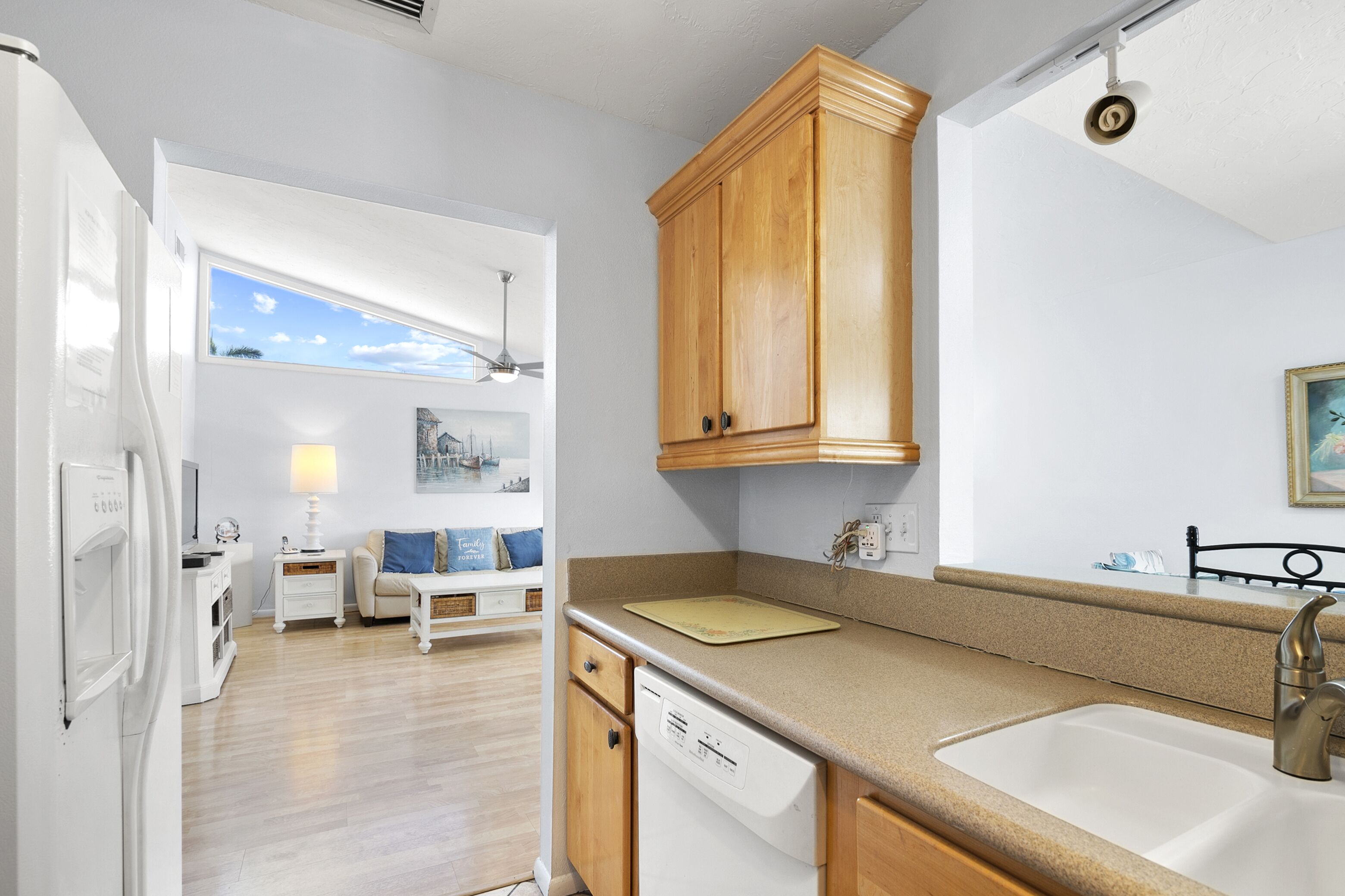 A compact kitchen with wooden cabinets, a built-in dishwasher, a sink, and a window overlooking a living area with a sofa and a lamp.