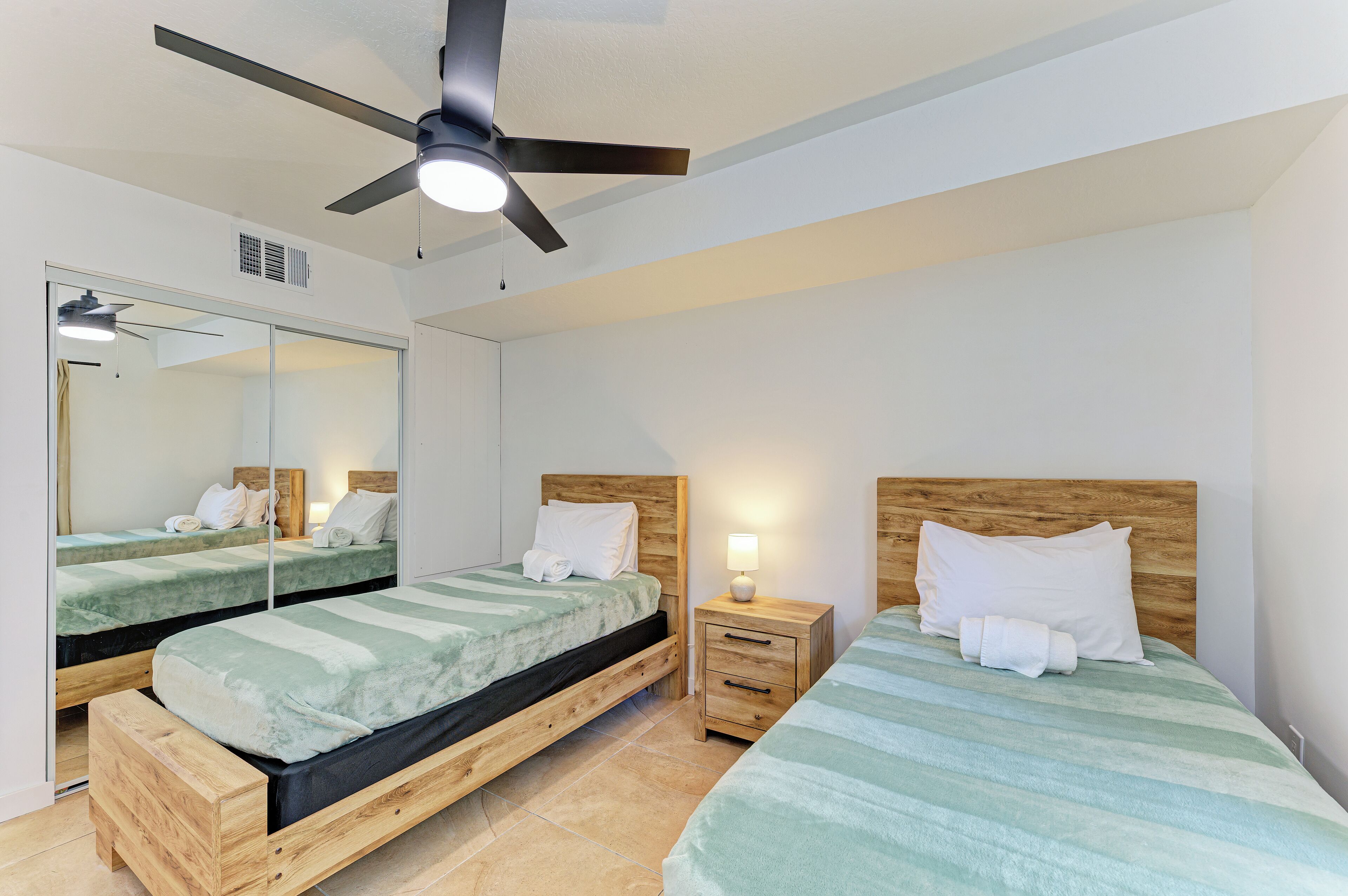 A hotel room with two single beds, wooden headboards, a ceiling fan, a bedside table with a lamp, and a large mirror.
