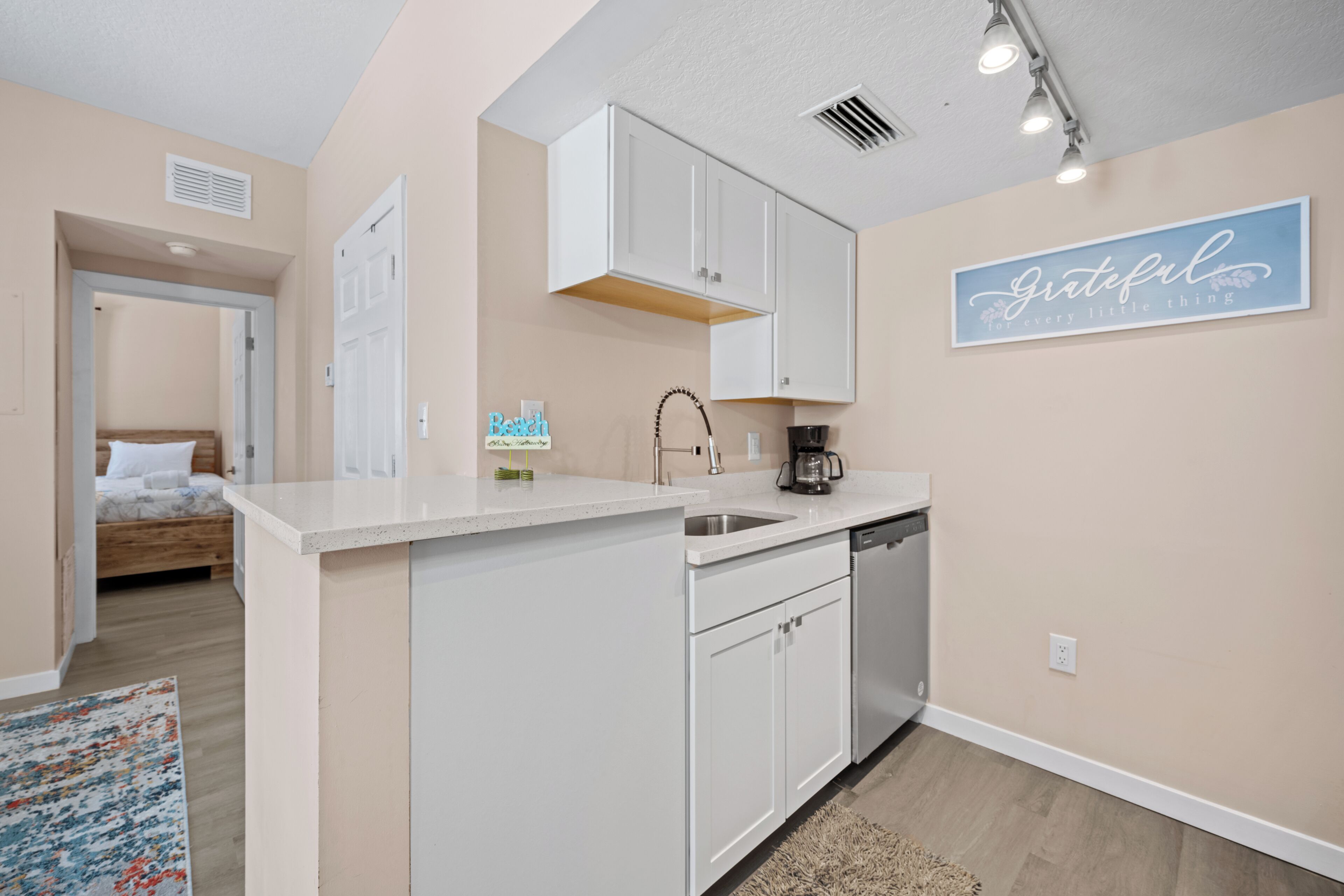 A compact kitchen with white cabinets, a coffee maker, and a sink. A sign on the wall reads "Grateful for every little thing."