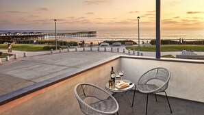 Een balkon met twee rieten stoelen, een tafel met een fles en glazen, en uitzicht op de oceaan en de pier.