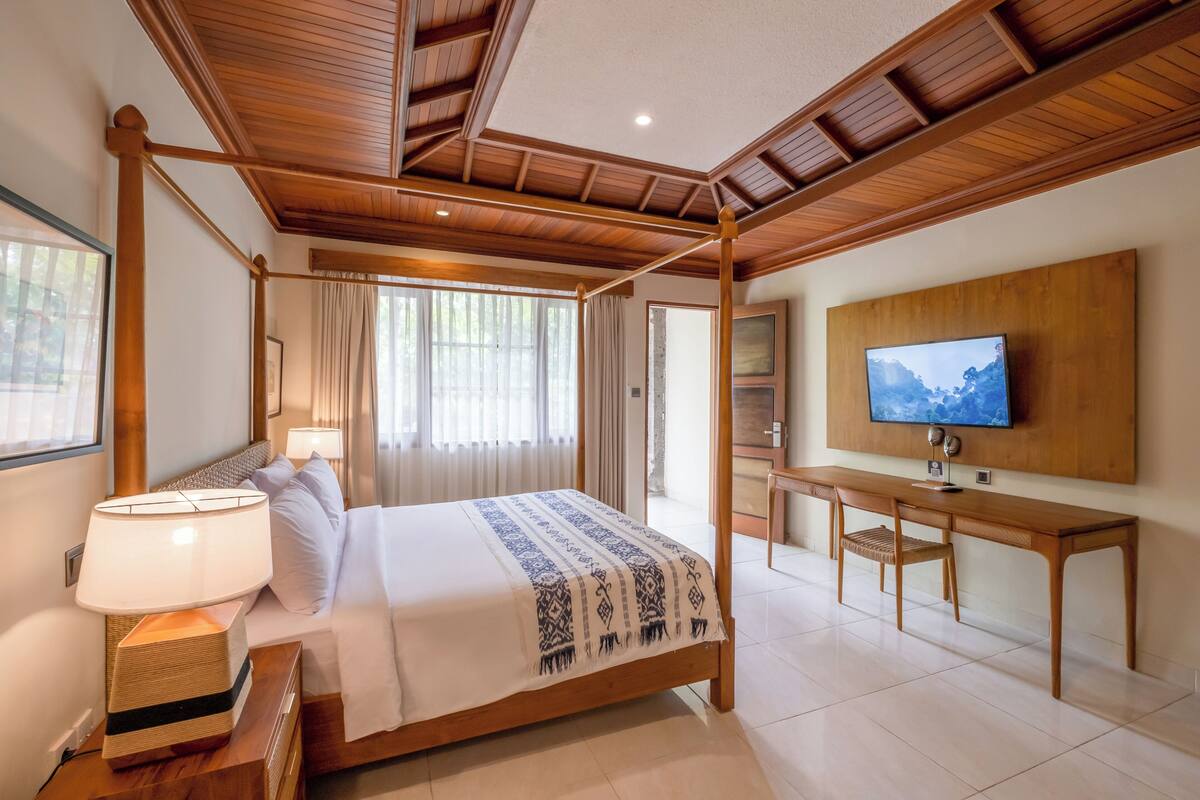 A hotel room with a four-poster bed, a desk with a chair, a television, and a large window with curtains.