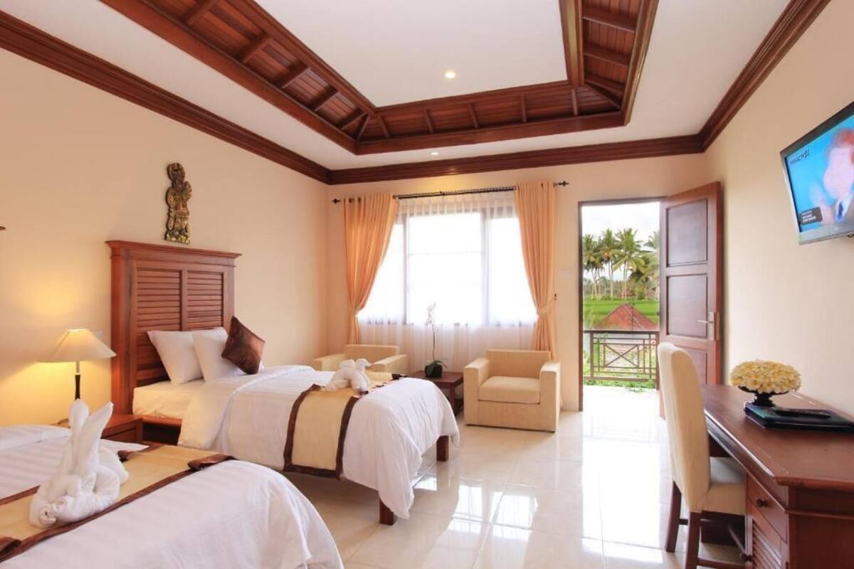 A hotel room with two beds, a desk, a chair, a TV, and a balcony with a view of palm trees.