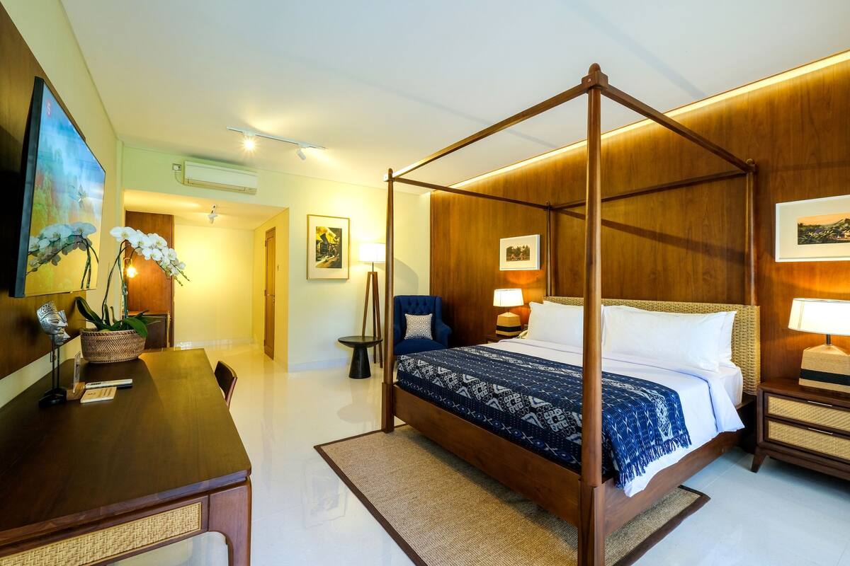 A four-poster bed with a patterned bedspread, a wooden nightstand with a lamp, a dining table with chairs, and a wall-mounted air conditioning unit.